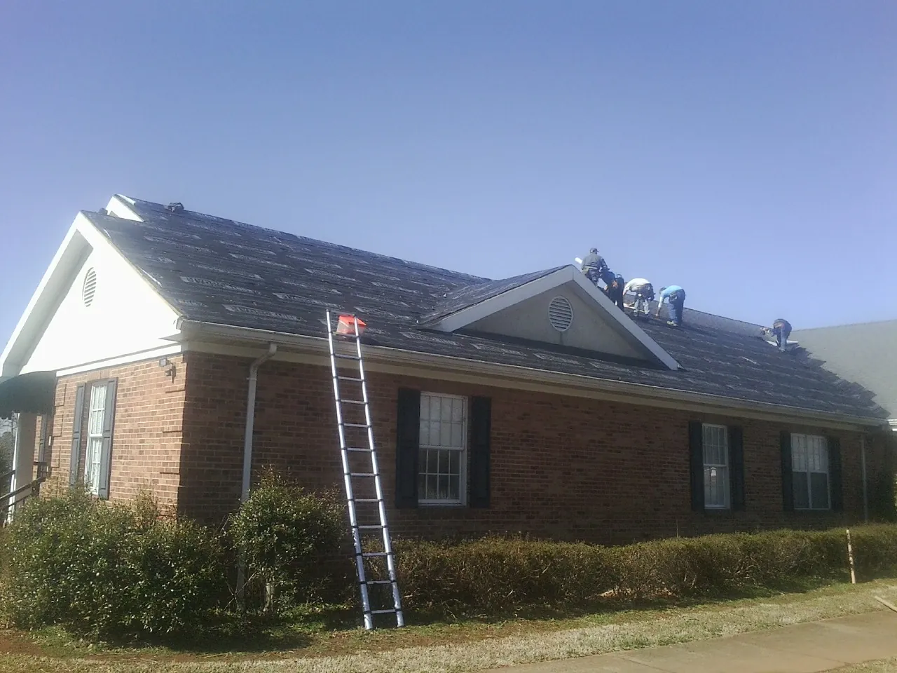 Crew tearing shingles off a brick ranch roof with a ladder set — Metal Roof Repair work by our crew in Haines City
